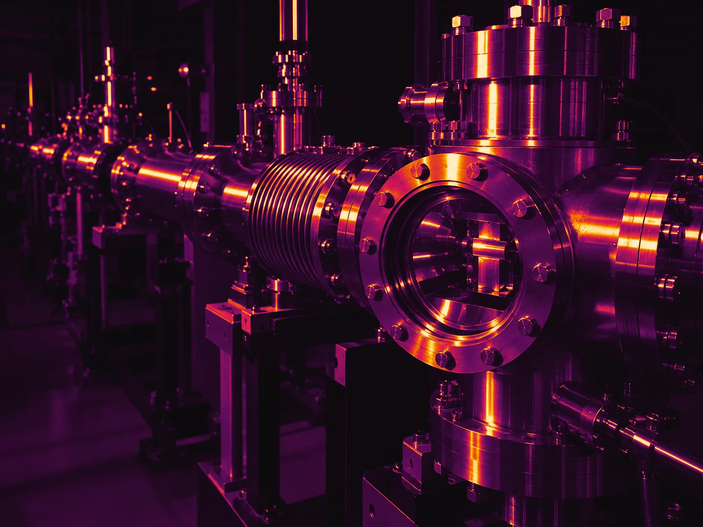 Illustrative photograph: a stainless accelerator beamline with a viewport target chamber, bellows and bolted flanges