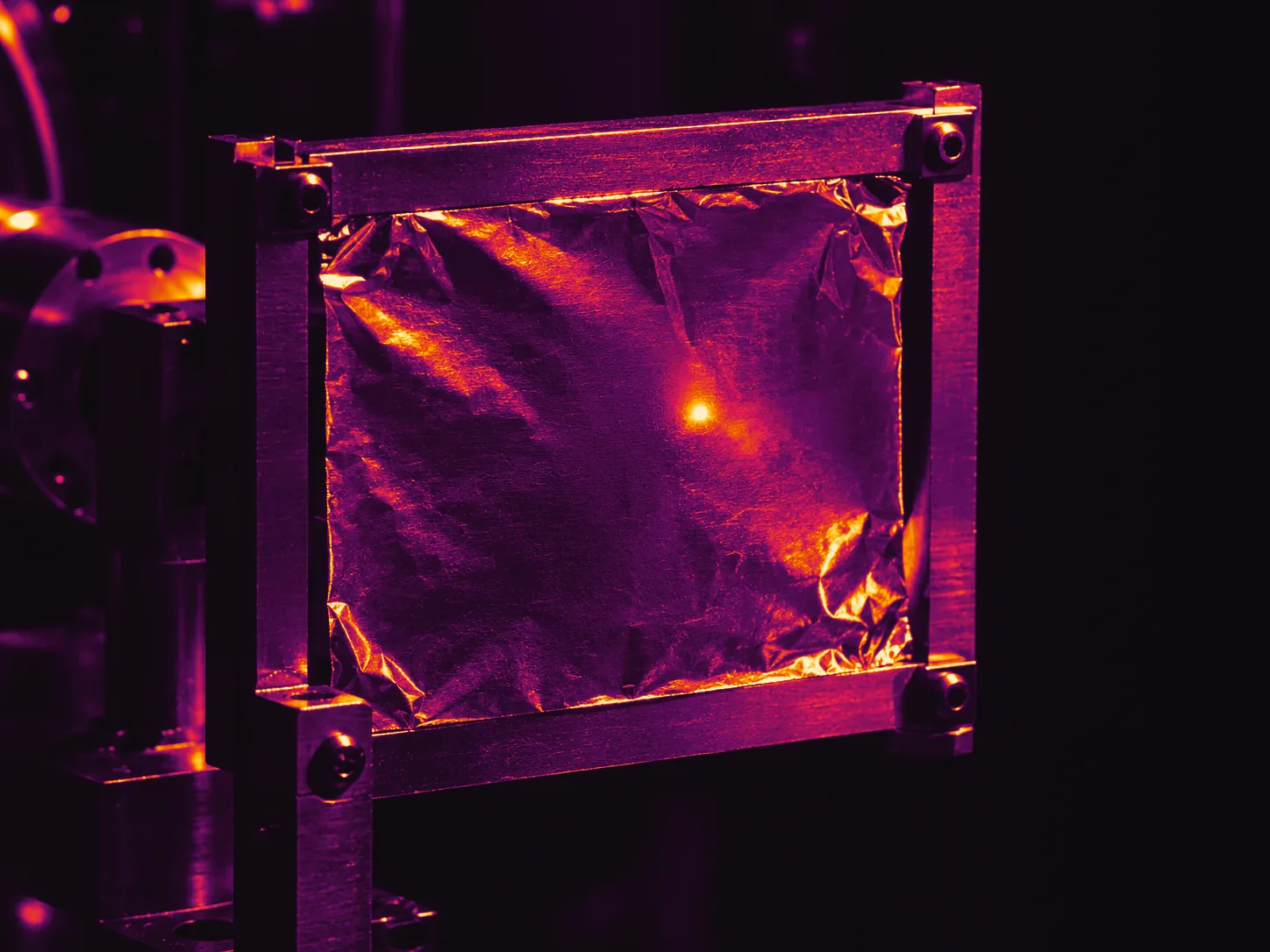 Illustrative photograph: a thin metal foil in a precise holder with a small bright impact spot